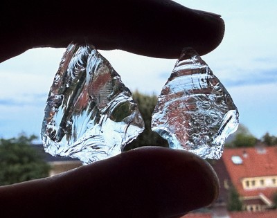 Foto vanaf een balkon. Een duim en een wijsvinger houden twee glazen pijlpunten tegen het licht. Ze hebben de vorm van een gitaar-plectrum, maar dan met een stammetje en een puntje. Je ziet goed hoe de breuklijnen van grote handelingen en kleine retouches lopen. Door de prisma werking, zie je in de pijlpunten de rode kleur van pannendaken, groen van de bomen, en blauw van de lucht.
