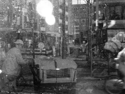 Another layered image. It is snowing, and it is shot from the inside of my workplace out. It's hard to tell what is snow and what is dirt on the window. A man with a cap pushes a cargo tricycle, a cyclists just leaves the frame to the right. Above it, the reflection of the camera in my hand. Modern buildings in the background and a huge flair. A selective ray of sunlight hits a traffic sign, and that sign is reflected onto the building across the street too.