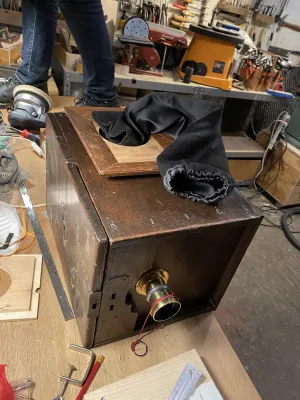 Camera is on it's side on the work bench. The door is getting finished, the hole for it is not there yet. The truck that allows for access to the camera, is draped on top of the door. It will be mounted into the round hole in the door. The feet and legs in the back are Pelle's.